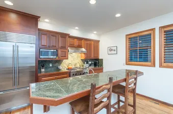 A fully equipped kitchen with wooden cabinetry, green tiled countertops, stainless steel appliances, and a breakfast bar with two wooden stools.